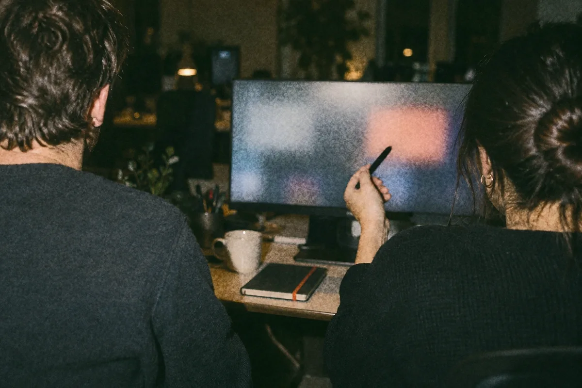 Two people seen from behind at a desk in the evening, one pointing at a large screen with a pen